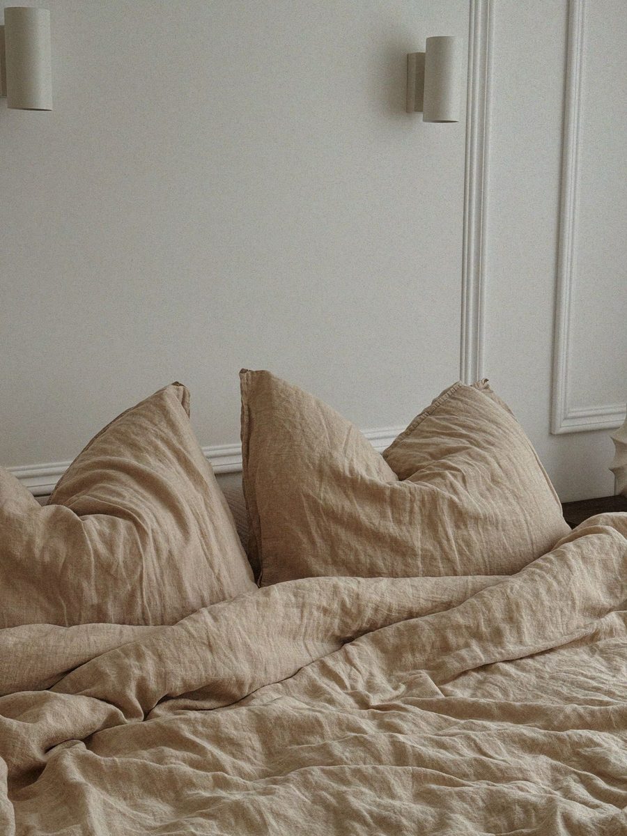 A bed made up in undyed oat linen, pillows stacked against a white wall